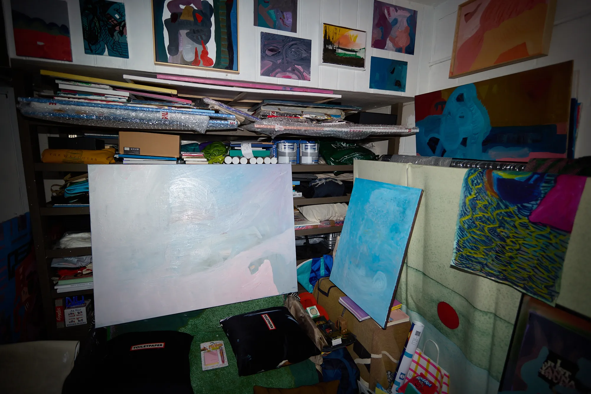 A shelf of small paintings above bundled canvases wrapped in bubble wrap and paint tins, with a large blue painting propped against the wall