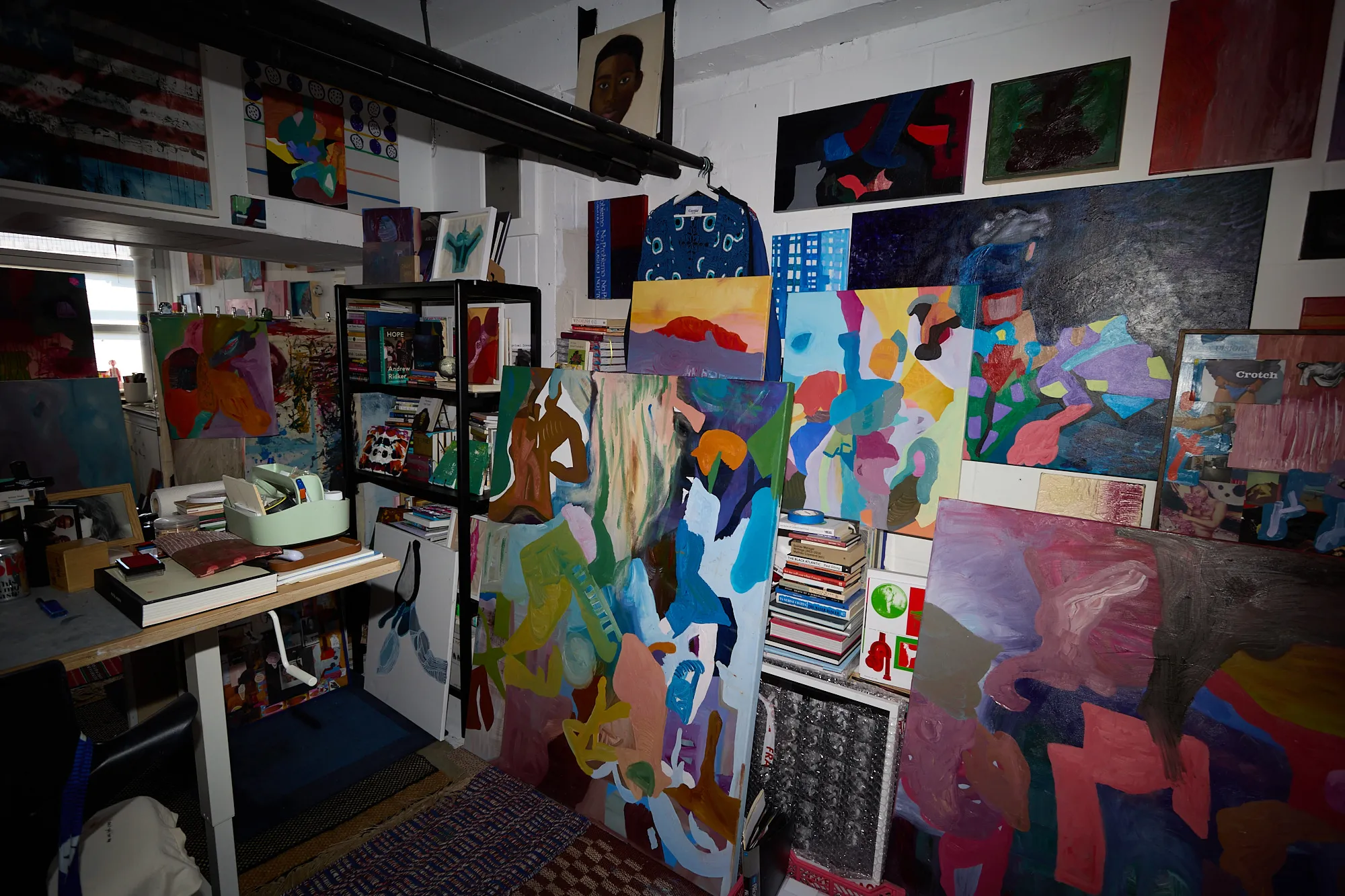 A dense corner of Omar Kholeif's studio, paintings stacked and hung everywhere, with the photographer caught as a translucent reflection in a mirror