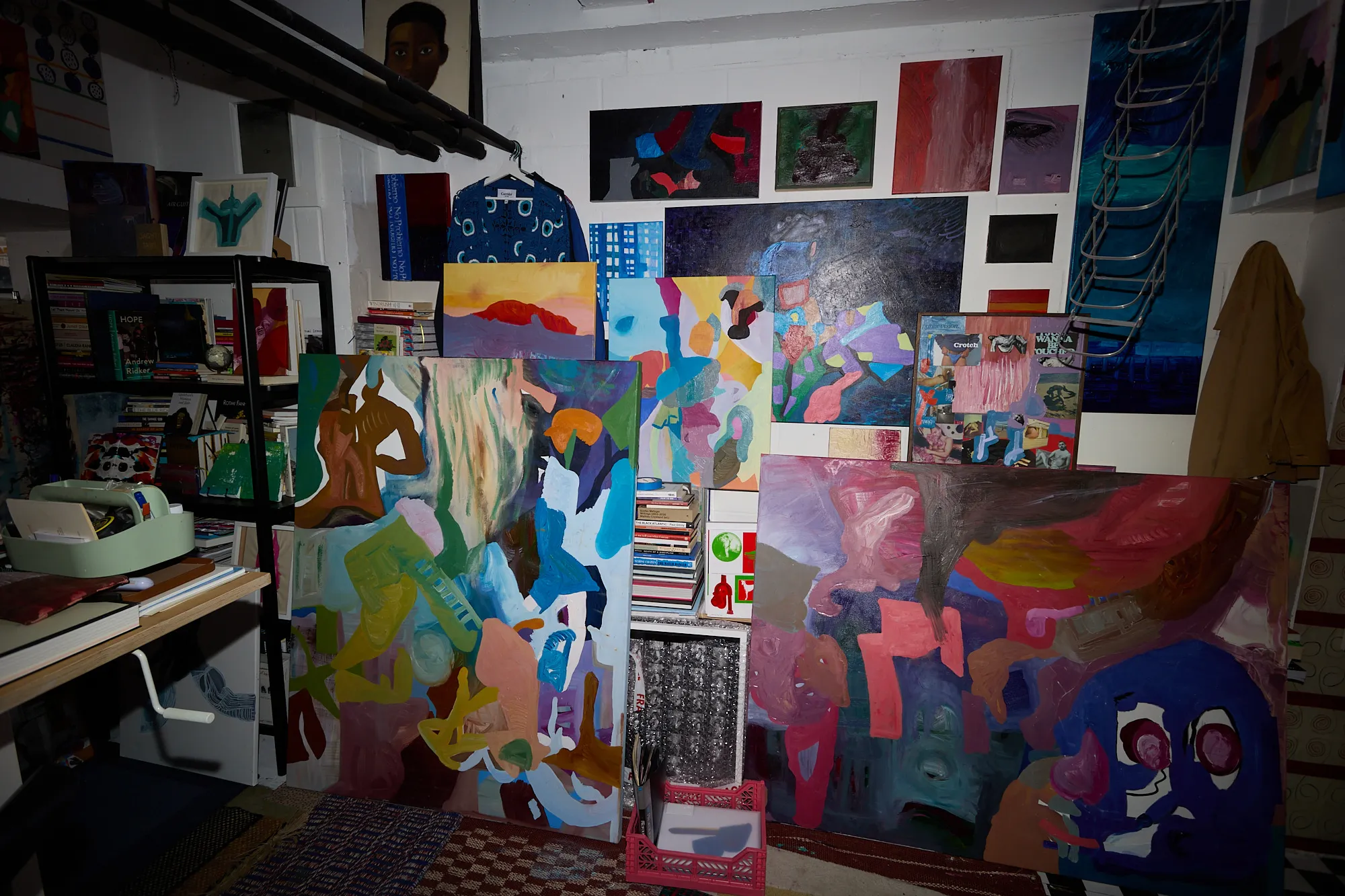 A dense cluster of paintings and a collage propped against the studio wall, beneath a ladder