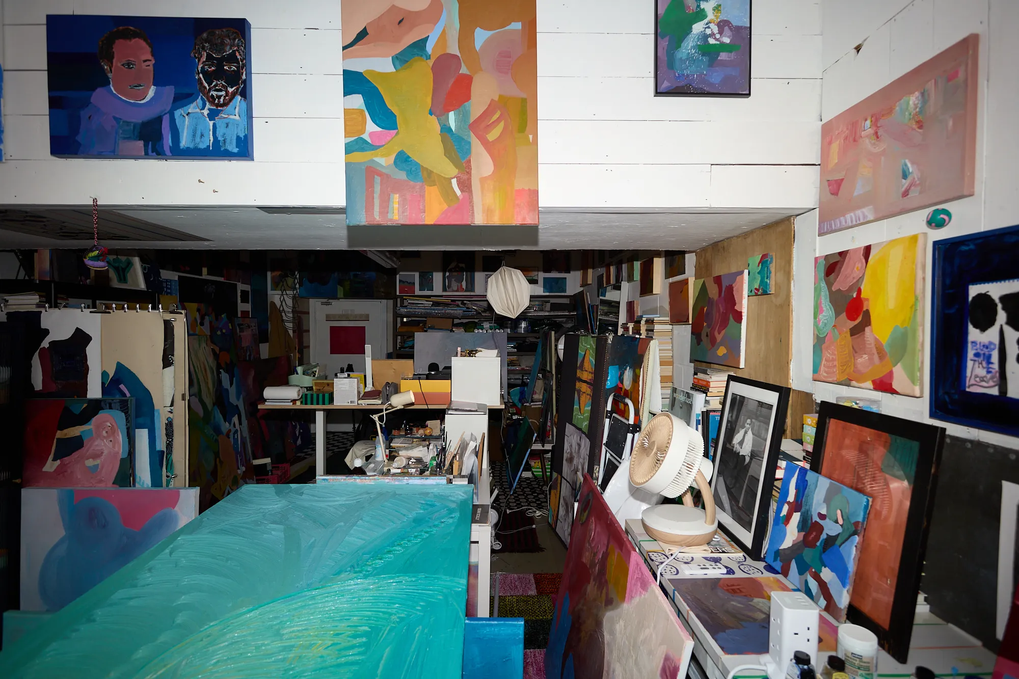A dense corner of Omar Kholeif's studio, paintings stacked and hung from every surface beneath a small watching portrait, before it was cleared