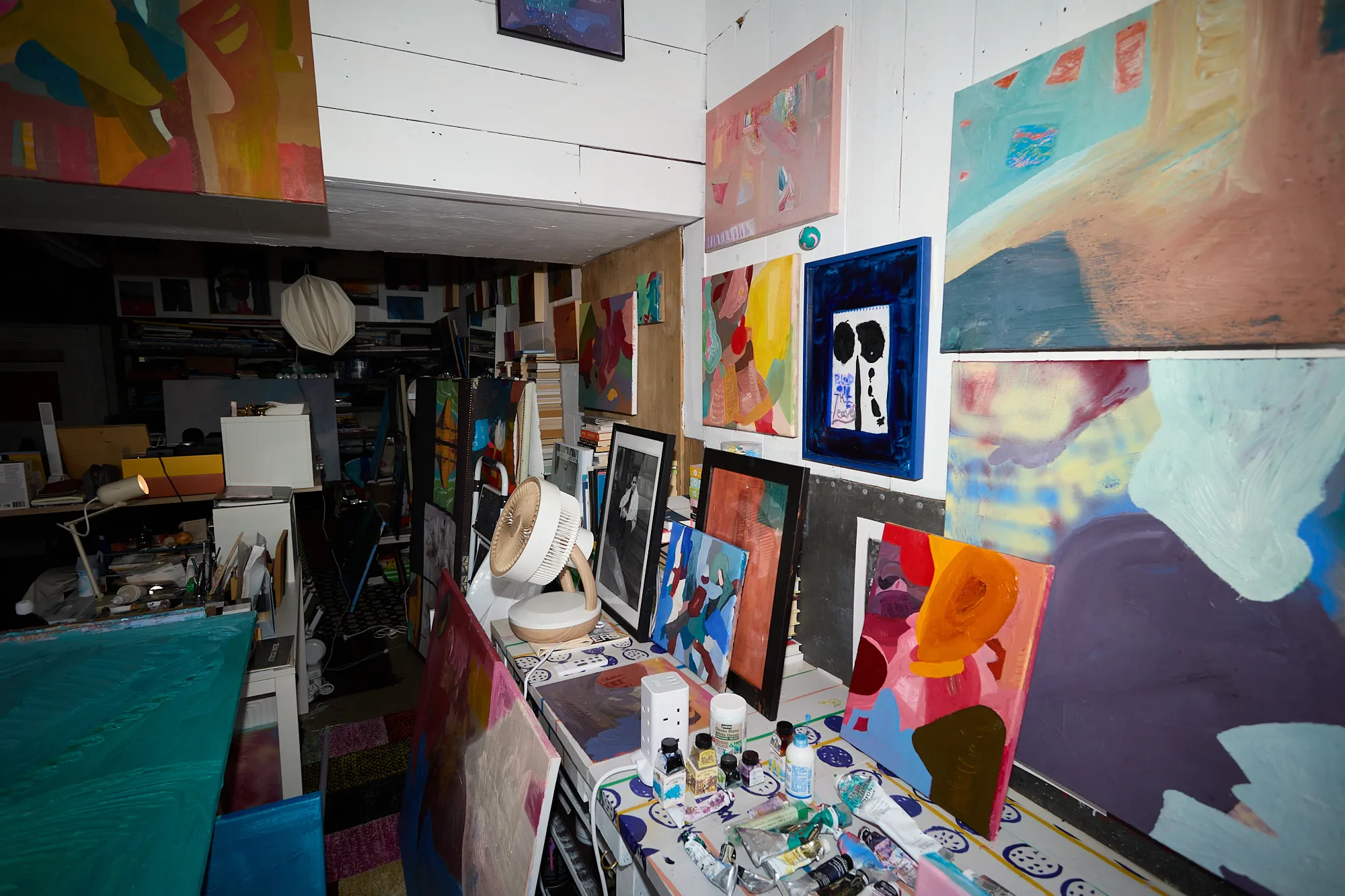A wall crowded with small and mid-sized paintings above a cluttered desk in Omar Kholeif's studio