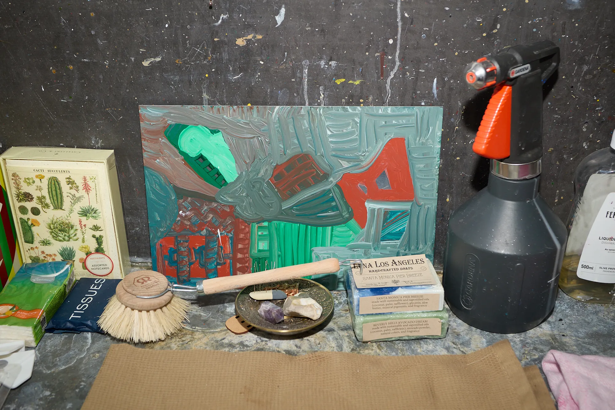 A small painting propped against a paint-stained wall, beside soaps, tissues and crystals near a sink in Omar Kholeif's studio