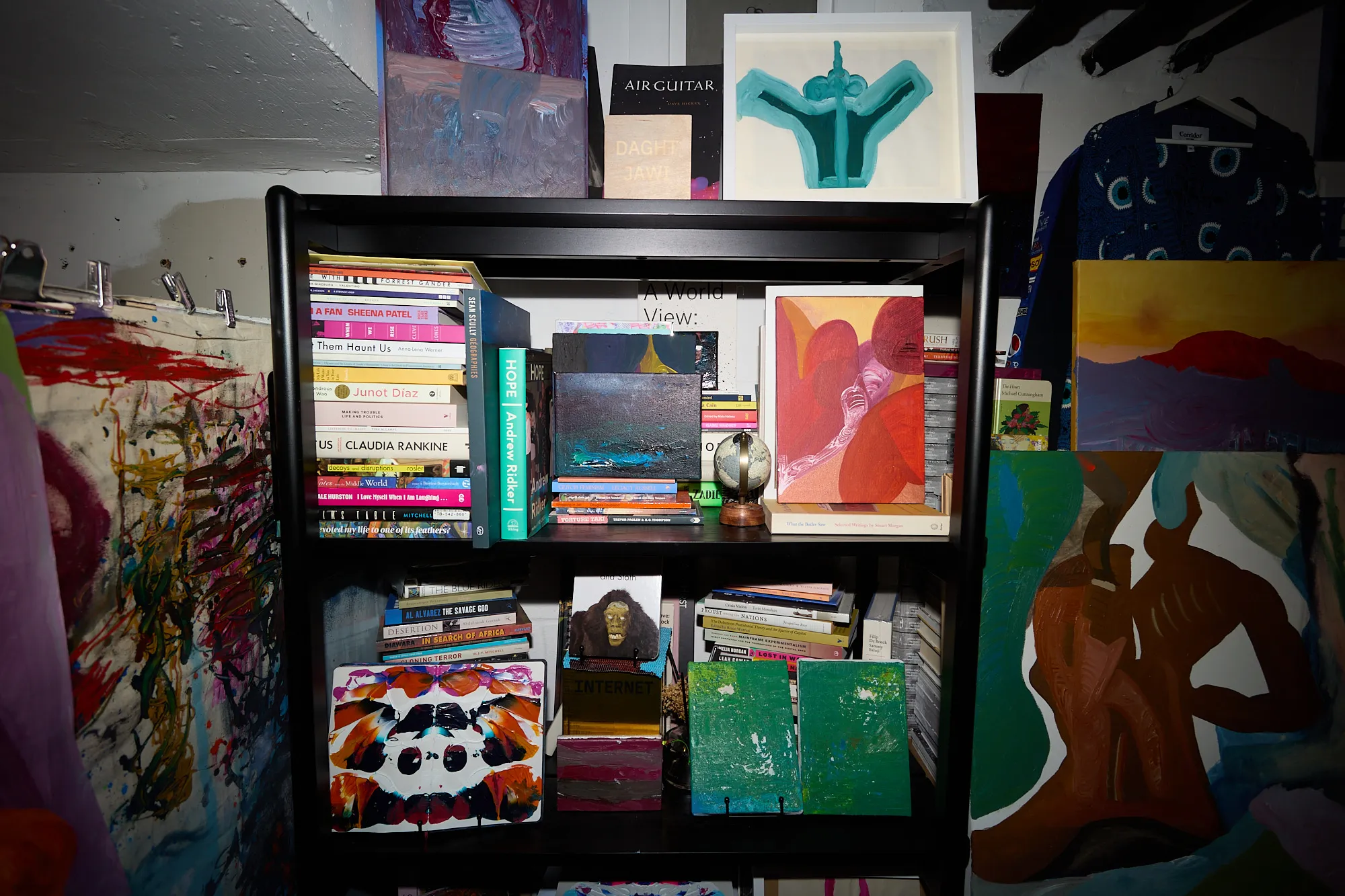 A bookshelf in Omar Kholeif's studio crowded with novels, art catalogues, small paintings and a globe