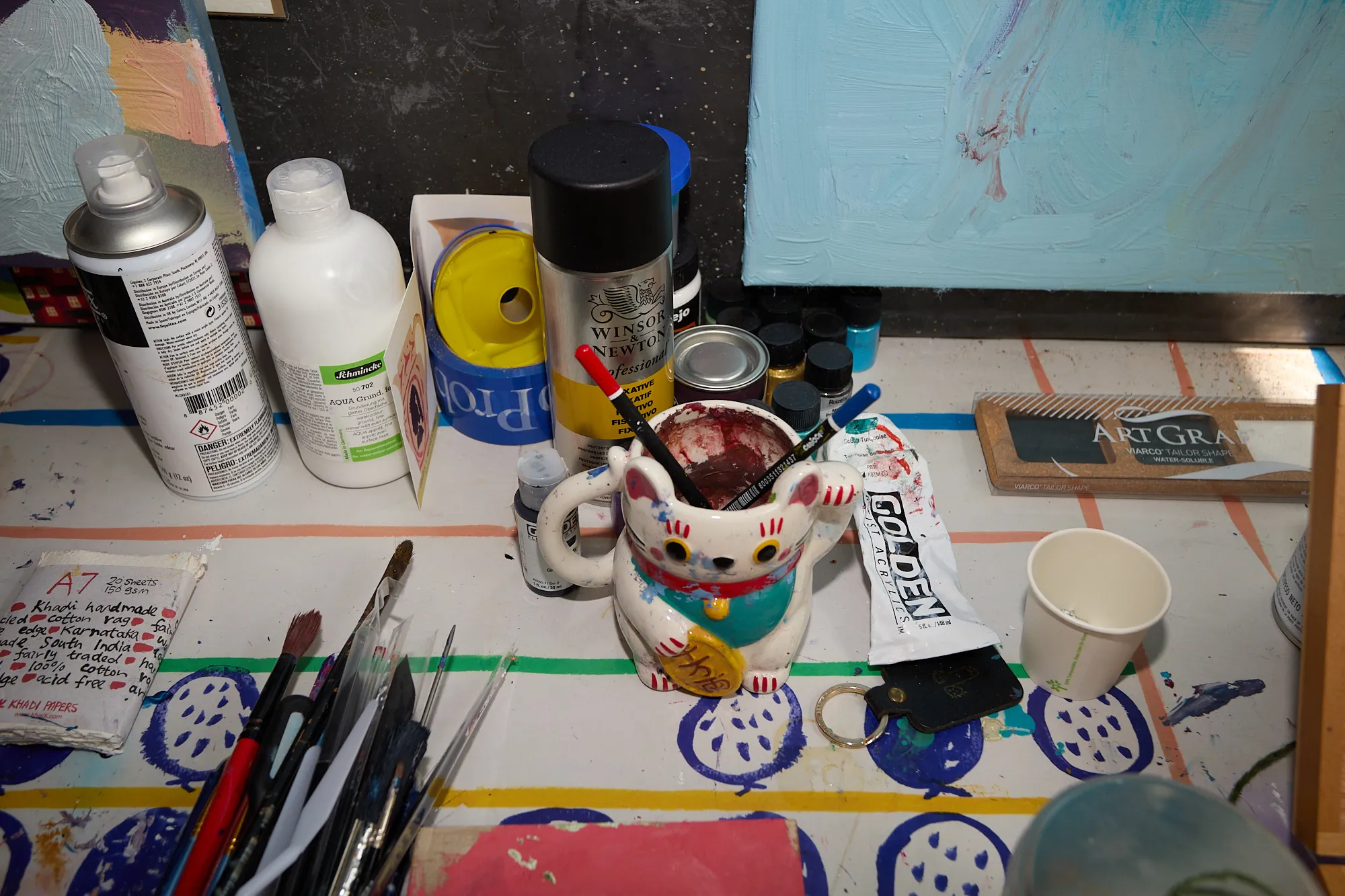 A lucky cat mug holding paintbrushes amid tubes of paint, keys and studio supplies on Omar Kholeif's desk