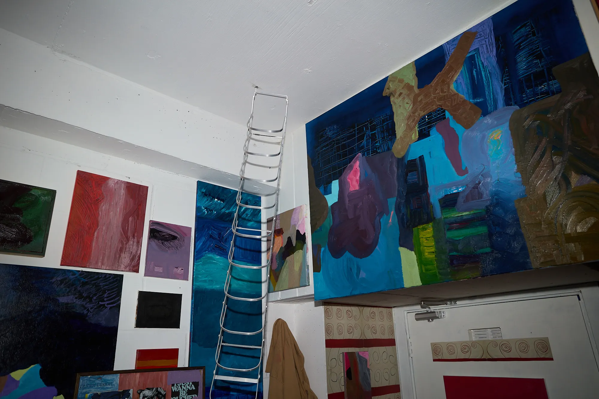 A ladder rising to a mezzanine beside a large blue painting, walls dense with more paintings in Omar Kholeif's studio