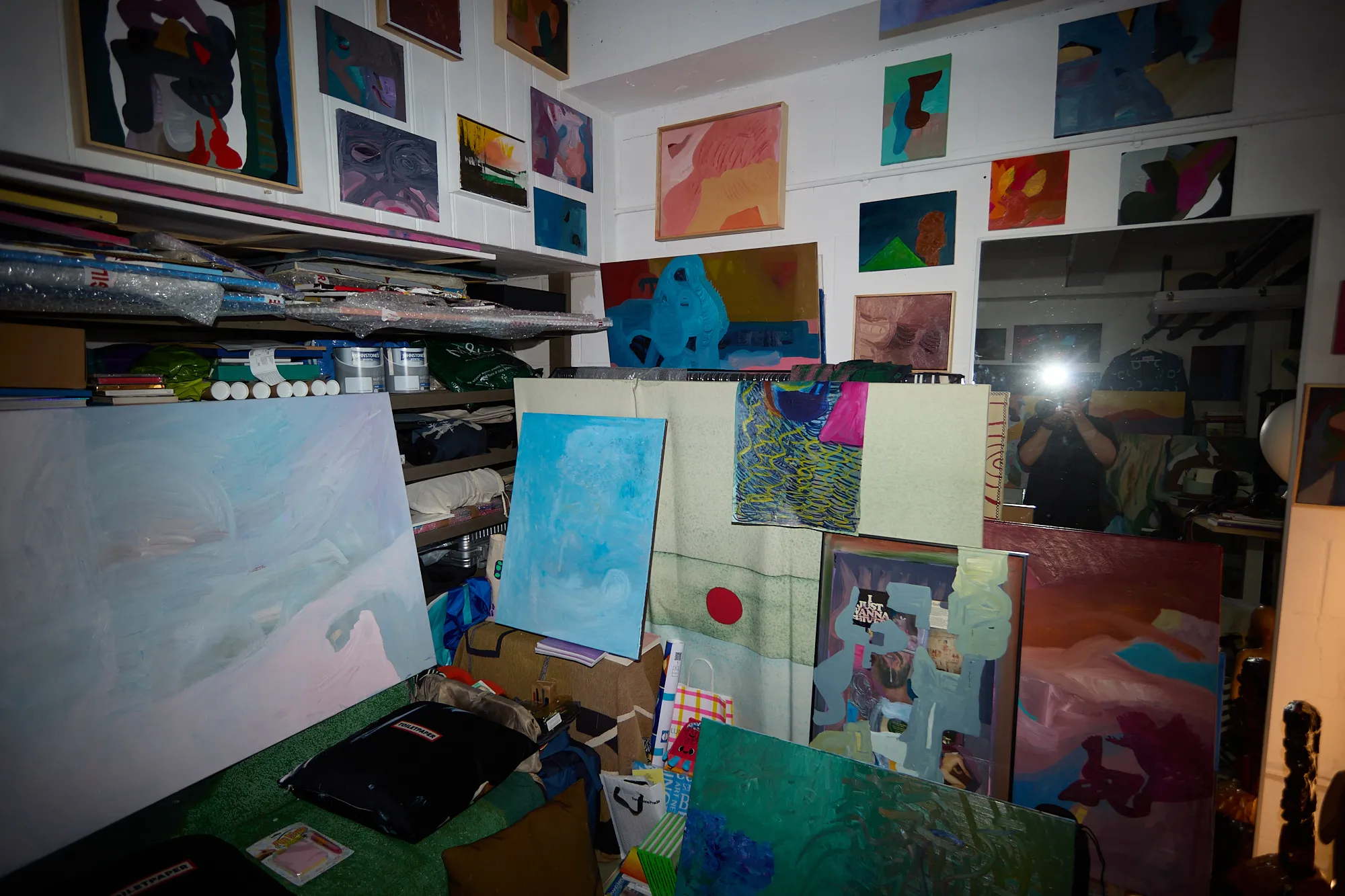 A dense corner of Omar Kholeif's studio, paintings stacked and hung everywhere, with the photographer's camera flash caught as a bright ghostly reflection in a mirror